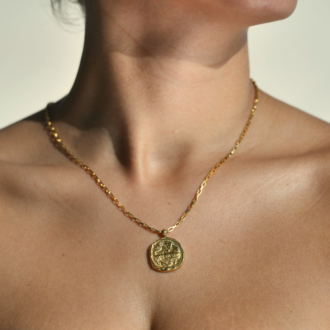 AQUARIUS Zodiac Medallion Gold Necklace. Irregular round medallion with an aquarius symbol in the middle. A vase throwing water in the middle of the medallion. The gold medallion is droped in a chain necklace.