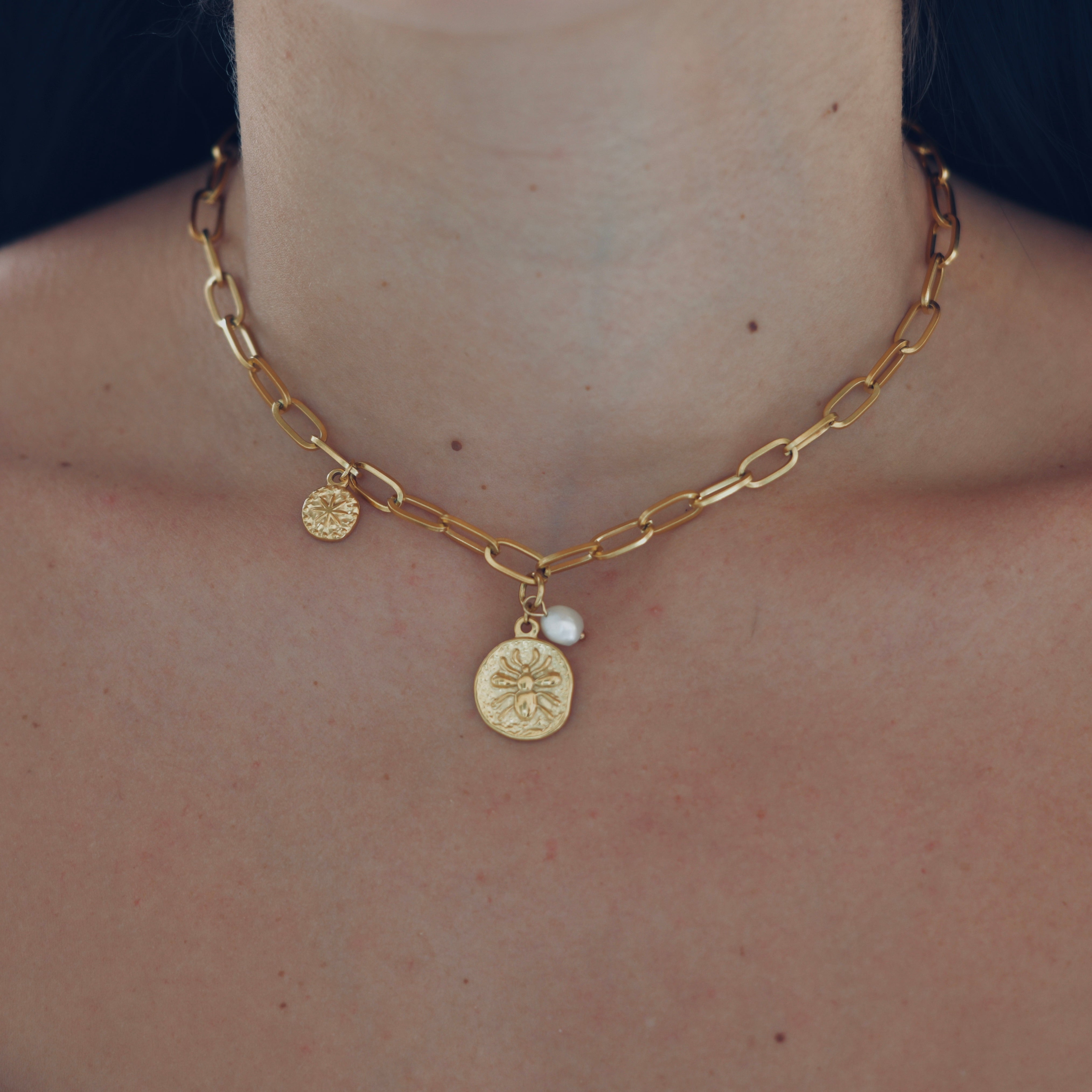 a medallion with a vintage texture. with a bee engraved on the medallion. a white pearl hangs on the side of the locket, on the other side hangs a smaller locket with a star engraved on it. the three parts are hanging on a gold chain.