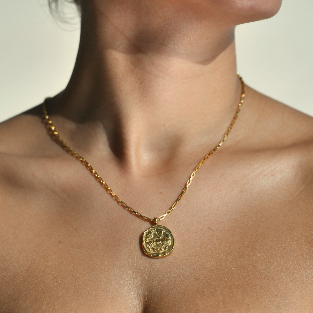    AQUARIUS Zodiac Medallion Gold Necklace. Irregular round medallion with an aquarius symbol in the middle. A vase throwing water in the middle of the medallion. The gold medallion is droped in a chain necklace. 