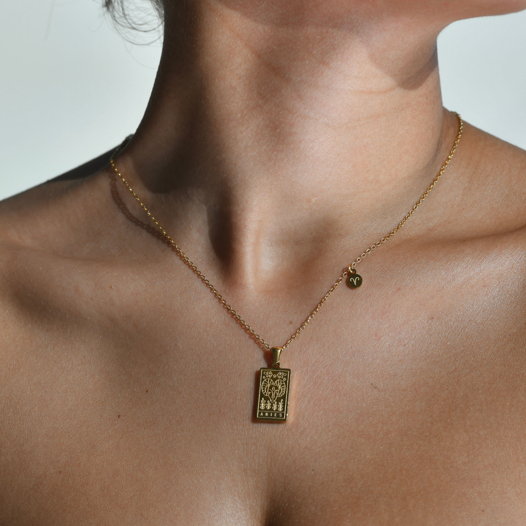 ARIES Zodiac Pendant Gold Necklace -The pendant is in a vertical rectangular shape. With the figure of the ram carved into it. The word aries is engraved at the bottom of the pendant. The pendant is gold and the chain on which the pendant hangs. In the corner of the chain there is a small round medallion with the ram symbol.