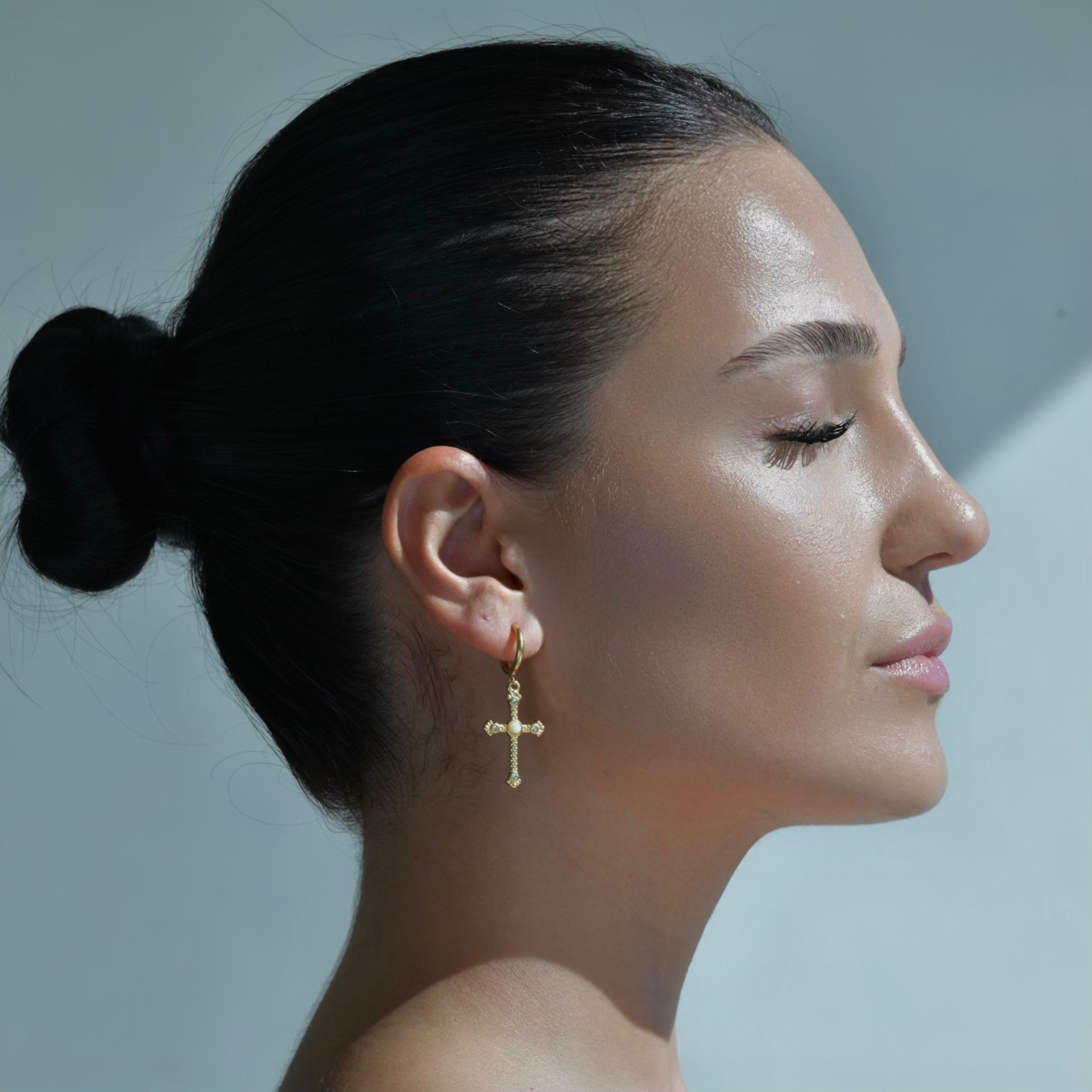 model wearing gold Earrings, A cross pendant on a small hoop. The cross is adorned by zircons and a white central white small pearl.