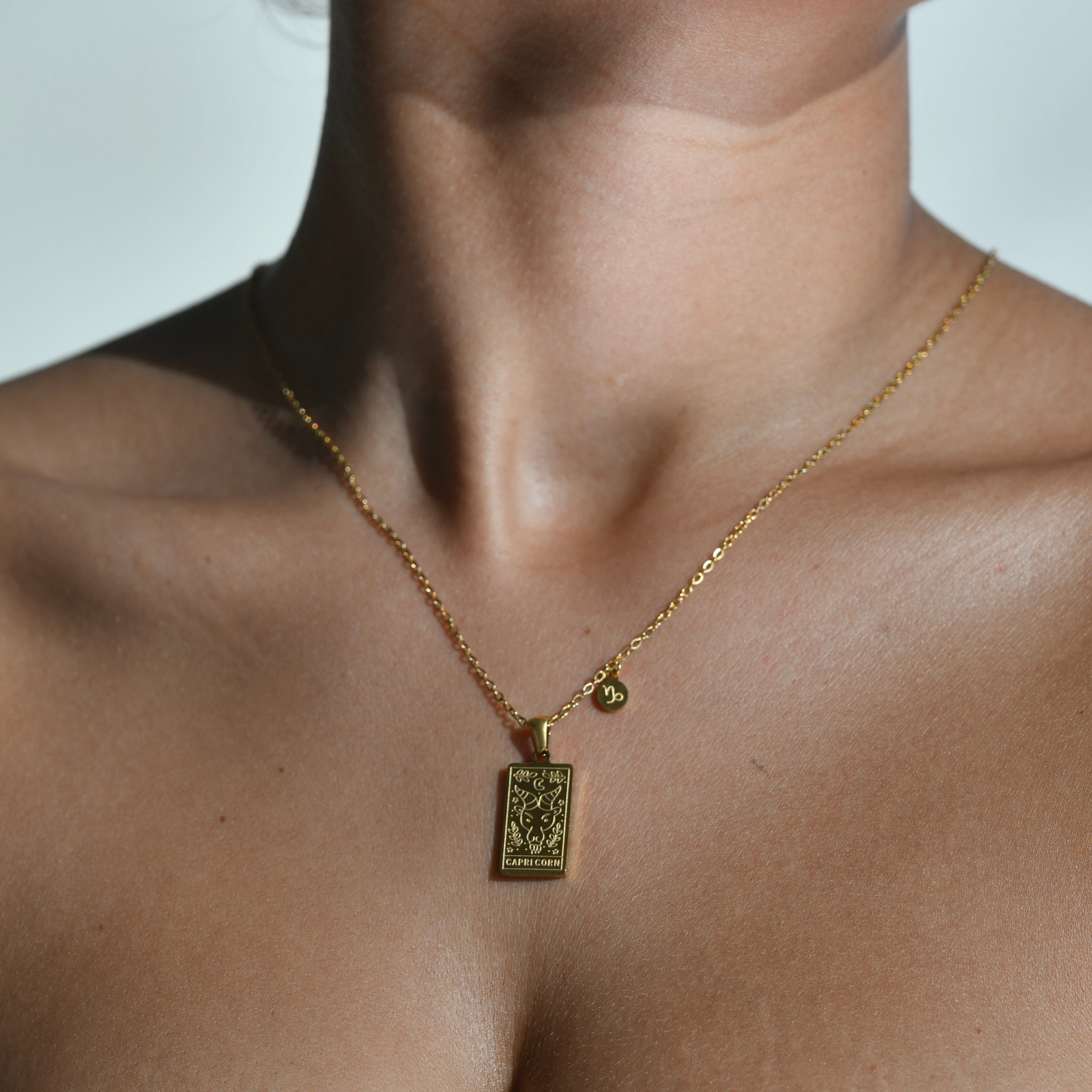 CAPRICORN Zodiac Pendant Gold Necklace. rectangular gold pendant. gold chain. the zodiacal sign of the goat with the goat's head is engraved on the pendant and the word capricorn is engraved on the bottom. at the side of the pendant attached on the chain is a small medallion with the capricorn symbol engraved on it.