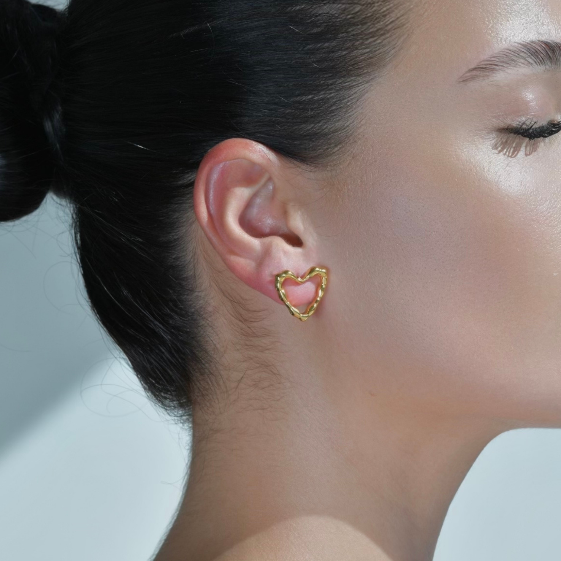 Woman wearing a Heart shape gold stud earrings. Irregular surface on the border of the heart shape.