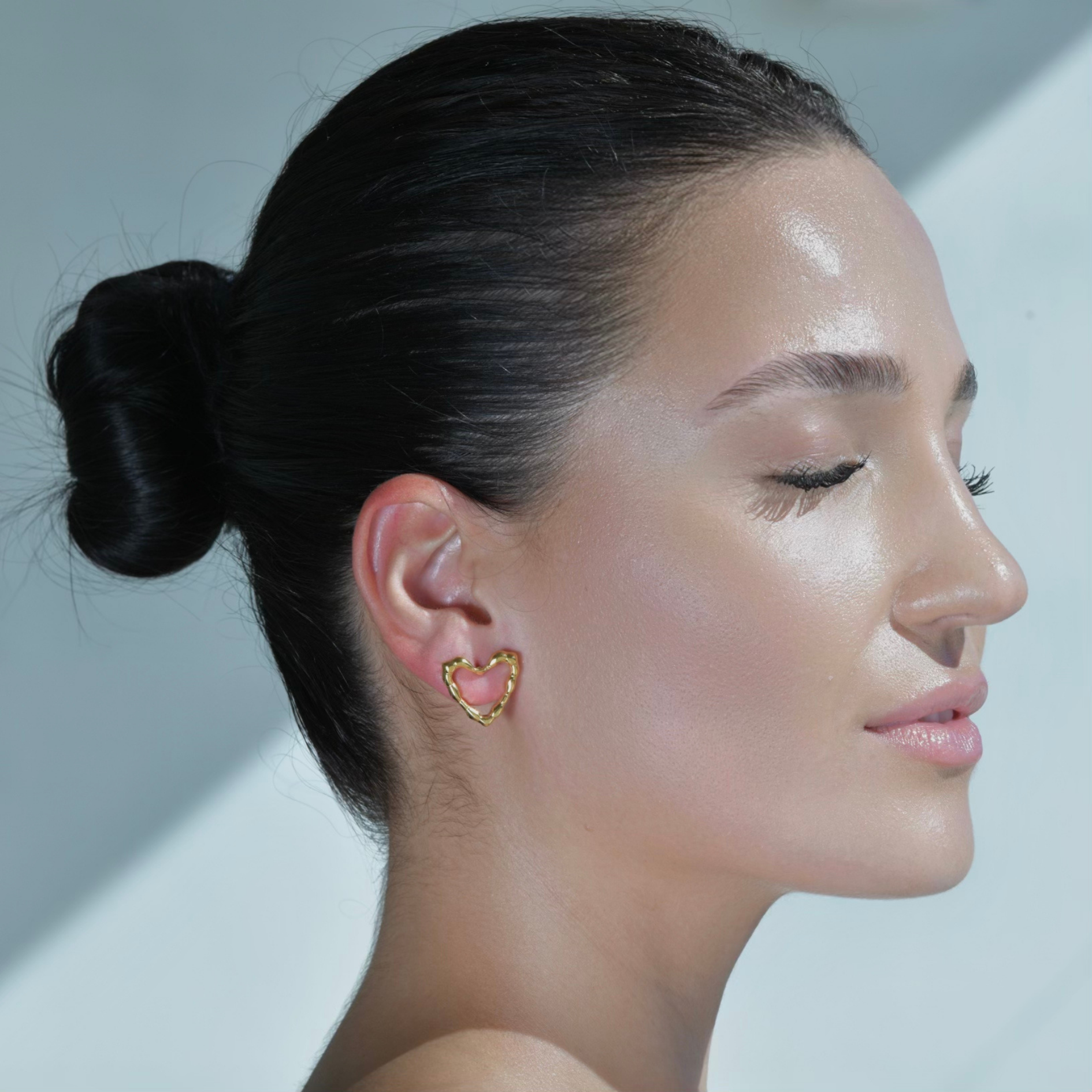 Woman wearing a Heart shape gold stud earrings. Irregular surface on the border of the heart shape.