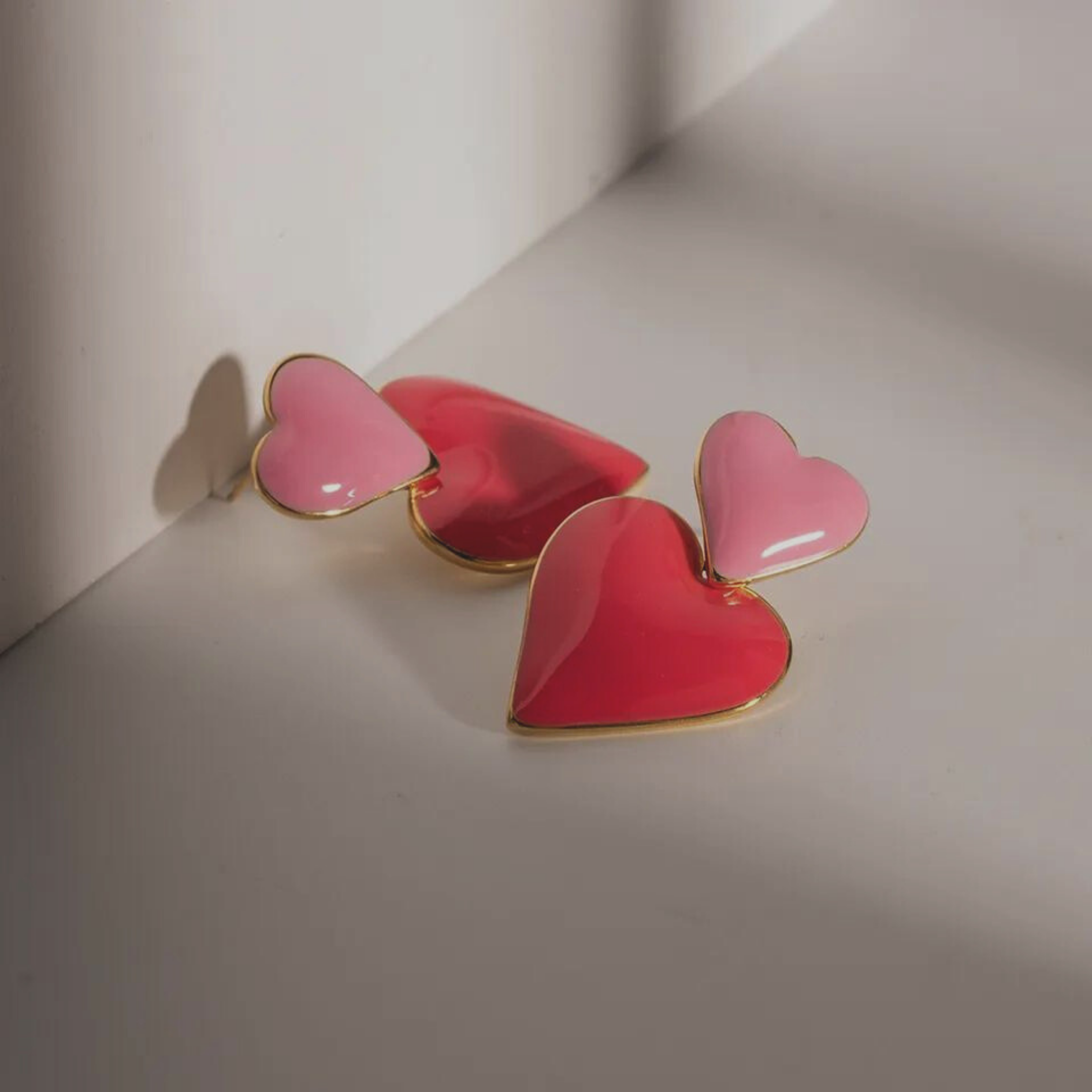 Gold Hearts Drop Earrings. Two hearts attched with one another, one painted in enamel baby Rose in the top and a bigger one Painted in pink enamel droping in the bottom.