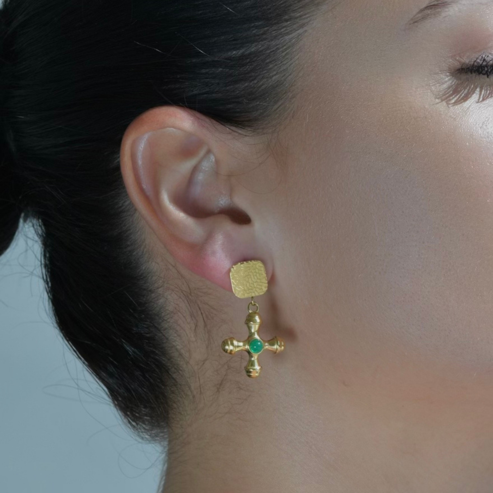 Gold Drop pair of earrings. The stud upper part has the shape of a square with oval corners and irregular texture.The drop part has the shape of a cross with a green stone in the middle of the cross. Taormina Gold Cross Earrings
